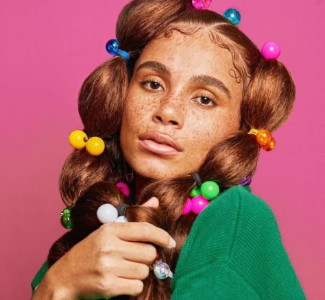 Light-skinned Black woman with colorful barrettes and vintage hairstyle, wearing bright clothing, gazing directly at the camera — symbolizing childlike freedom and inner peace